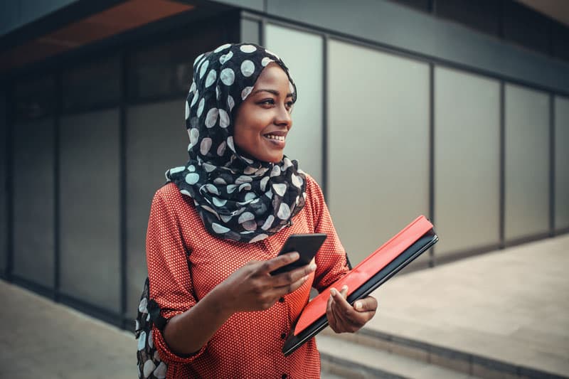 A smiling woman outside an office with a folder and a phone.