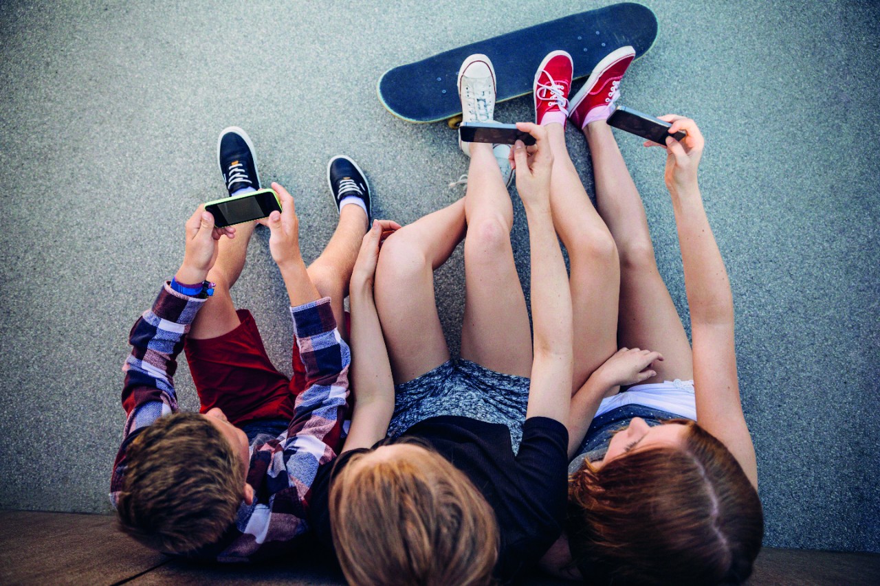 Three teenagers sitting outdoors with smartphones and skateboard