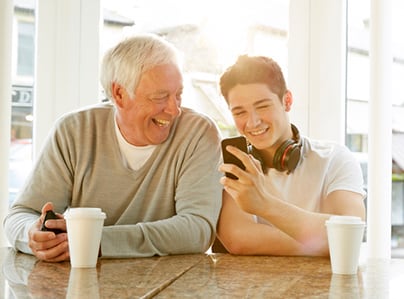 An elderly man with white hair and a young man with headphones around his neck are sitting at a café table, smiling and laughing as they look at a smartphone. Both have coffee cups in front of them. The scene is brightly lit by large windows in the background, creating a warm and cheerful atmosphere.