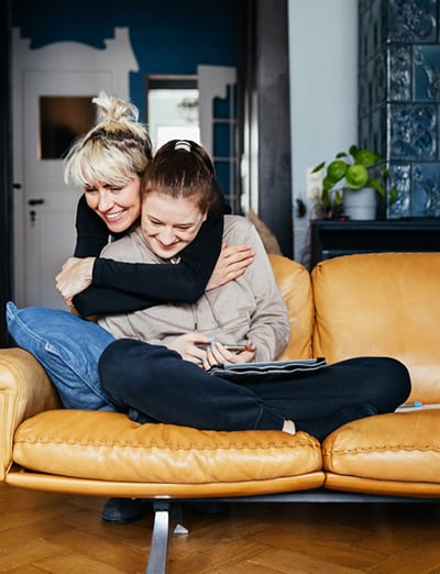 A mother and daughter sit on a yellow leather couch, sharing a warm hug and smiling. The mother, with blonde hair in a bun, wraps her arms around her daughter, who has brown hair tied back. They are in a cozy living room with blue and white decor and wooden flooring. The moment captures love and togetherne