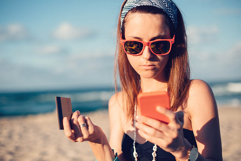 A girl on a beach looking at her phone and holding a credit card.