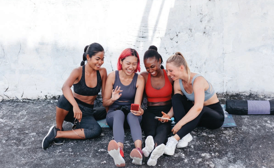 Four sporty women sitting on the ground and smiling and waving at a phone screen.