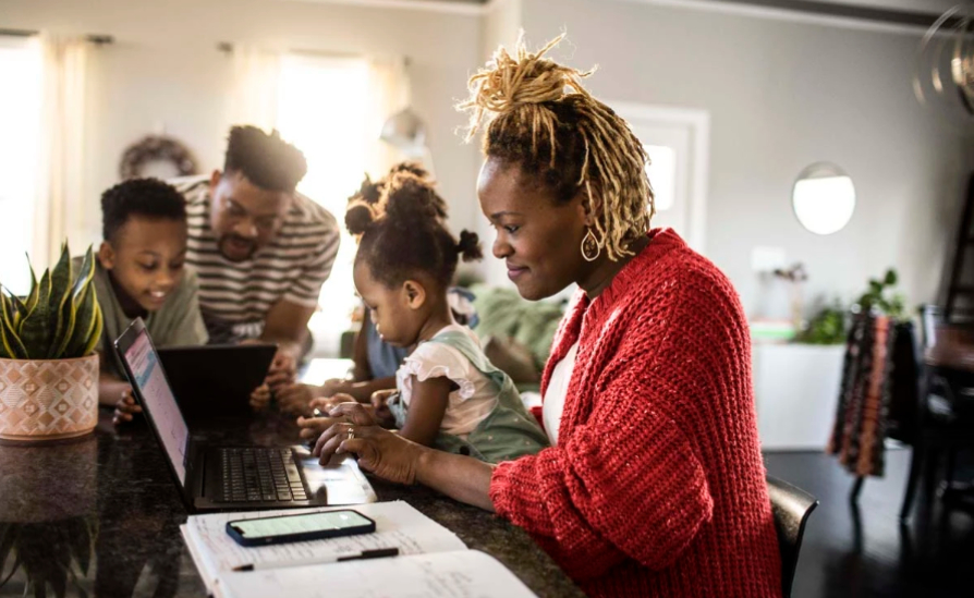 A family around a kitchen table where the Mum is working on a laptop and smiling.