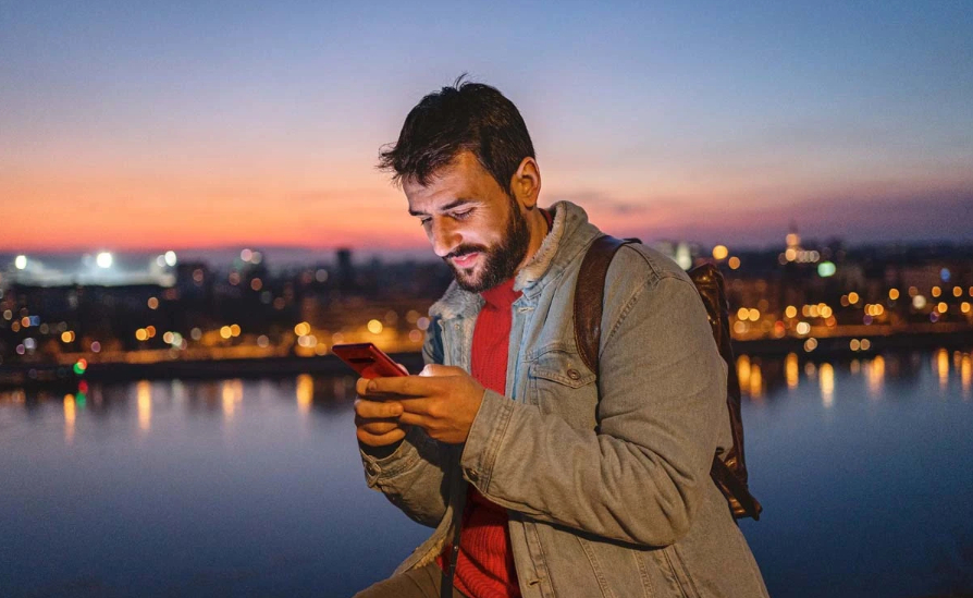 A man in front of a cityscape and river at dusk, looking at his phone.