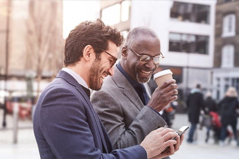Two men on street laughing and looking at phone screen.